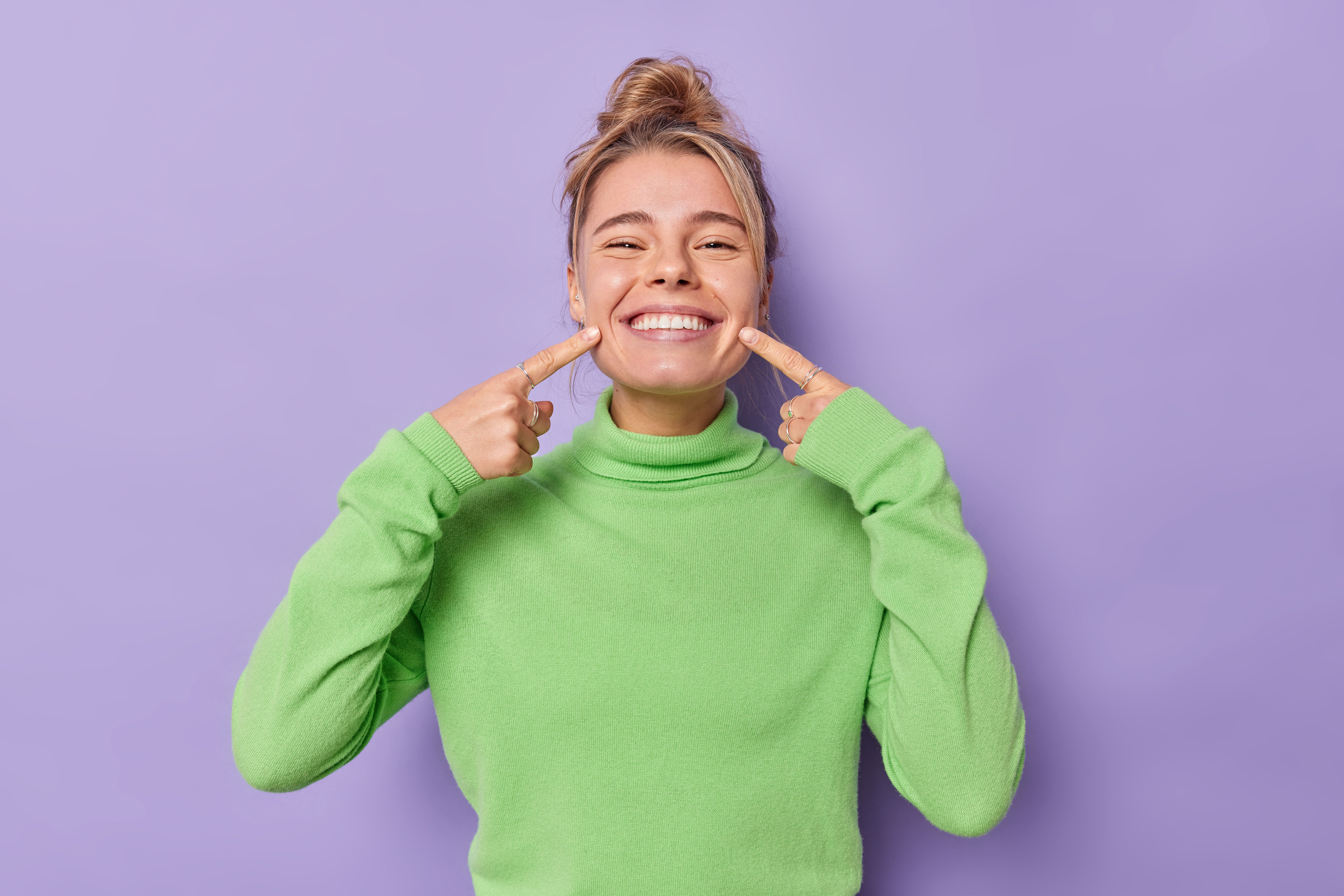 beautiful-happy-fair-haired-european-woman-points-index-fingers-mouth-forces-cheerful-smile-shows-perfect-white-teeth-wears-green-jumper-being-good-mood-isolated-purple-background.jpg